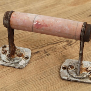 Puede incluir: Un portarrollos de papel higiénico antiguo con aspecto envejecido. El rodillo central es de color rosa descolorido, sostenido por soportes y placas de montaje de metal envejecido. El metal muestra signos de óxido y pintura descascarillada, dándole una estética vintage.