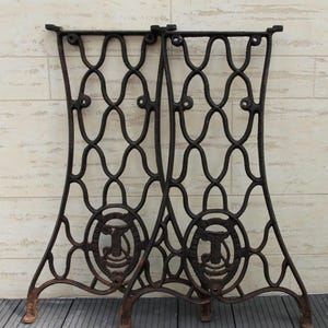 May include: Pair of antique cast iron sewing machine legs. The legs are dark brown with an ornate, openwork design featuring oval and circular patterns. Each leg has a circular emblem with the letter "T".
