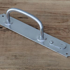 Könnte beinhalten: Ein silberfarbener Türgriff aus Metall mit einem gebogenen Bügel und einer rechteckigen Grundplatte. Die Grundplatte hat abgerundete Kanten und Schraubenlöcher. Der Griff ist zum Ziehen konzipiert. Die Hardware ist wahrscheinlich für eine Tür oder ein Tor. Ca. 20 cm lang.