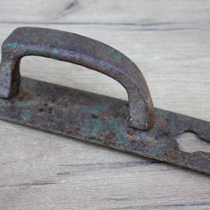 May include: An antique door handle with a curved grip and a rectangular backplate. The handle and backplate are made of aged metal with a rusty brown color and hints of green patina. The backplate has a keyhole.