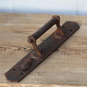 Może przedstawiać: Rustykalna klamka do drzwi w stylu antycznym, wykonana z ciemnego, zwietrzałego metalu. Klamka posiada długą, prostokątną płytkę tylną z ozdobnym elementem w kształcie rombu i cylindrycznym uchwytem. Metal ma postarzany wygląd.