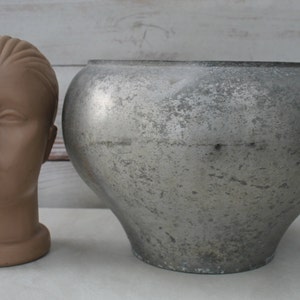 Puede incluir: Una olla grande, redonda, de metal de color plateado con una superficie texturizada. La olla está sentada sobre una superficie blanca junto a una cabeza de maniquí marrón.