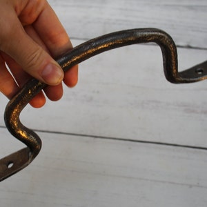 May include: A rustic, dark brown metal handle with a curved design. The handle has a textured surface and two mounting plates with pre-drilled holes. The handle is held in a hand against a light wood background.
