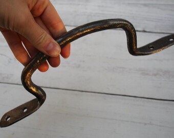 May include: A rustic, dark brown metal handle with a curved design. The handle has a textured surface and two mounting plates with pre-drilled holes. The handle is held in a hand against a light wood background.