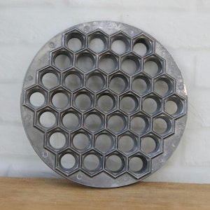 May include: A circular metal pelmeni maker featuring a repeating honeycomb pattern of hexagonal openings. The tool is constructed from a matte grey aluminum material and is designed with a flat outer rim, providing a structured grid for shaping multiple dumplings simultaneously in a kitchen setting.