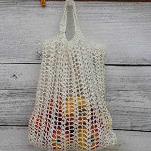 Könnte beinhalten: Eine weiße gehäkelte Netztasche mit Henkel, gefüllt mit orangefarbenen und gelben Früchten. Die Tasche hängt an einer weißen Holzoberfläche.