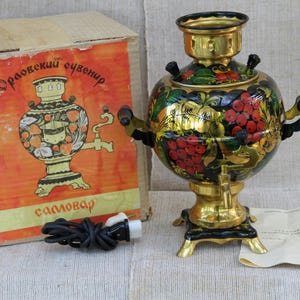 Può includere: Un samovar elettrico nero e oro con un motivo floreale e di bacche dipinto a mano, due manici laterali, un rubinetto e una base. Sono inclusi una scatola di cartone con il testo "Орловский сувенир" e "самовар", un cavo di alimentazione nero e un manuale cartaceo.