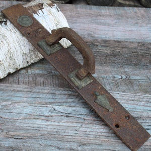 May include: A rusty metal door handle with a long, rectangular base and a curved handle. The handle is made of iron and has a dark brown patina. The base of the handle has several decorative elements, including a small, round knob and a larger, rectangular plate.