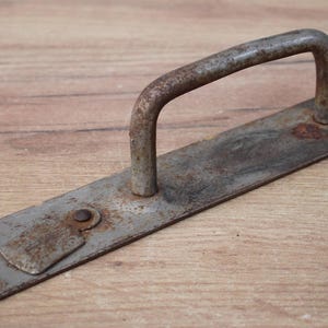 Könnte beinhalten: Ein rostiger Metalltürgriff mit einem dekorativen Design. Der Griff ist an einer Metallplatte mit abgerundeter Kante befestigt.