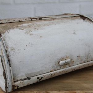 May include: An antique white bread box with a roll-top lid. The metal box shows signs of wear, with chipped paint and rust, giving it a vintage, rustic appearance. The box sits on a wooden surface.