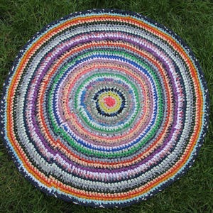 Peut inclure: Un tapis rond fait à la main avec un motif en spirale. Le tapis présente des cercles concentriques de différentes couleurs, notamment le rouge, l'orange, le jaune, le vert, le bleu, le violet et le gris. Le tapis est fait de bandes de tissu et a une bordure bleu foncé.