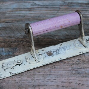 May include: A vintage white metal towel bar with a pink plastic handle. The handle is attached to the bar with metal brackets. The bar has a distressed, aged look.