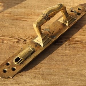 Puede incluir: Un tirador de puerta de latón antiguo con una placa trasera rectangular y un asa curvada. El tirador muestra signos de edad con un acabado deslustrado y algunas manchas oscuras. El tirador mide aproximadamente 25 cm de largo.