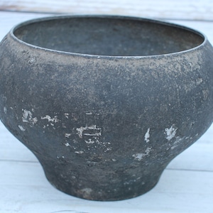 May include: A weathered, dark gray cast iron pot with a wide, open top and a rounded body. The pot has a slightly flared base and shows signs of wear, with areas of paint loss, giving it a rustic appearance.