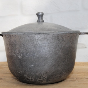 Puede incluir: Una olla de cocina vintage de hierro fundido con tapa. La olla es de color gris oscuro con una superficie texturizada y dos asas laterales. La tapa tiene un pequeño asa en la parte superior. La olla está sobre una superficie de madera.