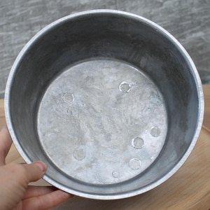 Könnte beinhalten: Ein silberner Aluminiumtopf mit einer glatten, glänzenden Oberfläche. Der Topf hat einen runden Boden und gerade Seiten. Am Boden des Topfes befinden sich mehrere kleine, runde Vertiefungen.