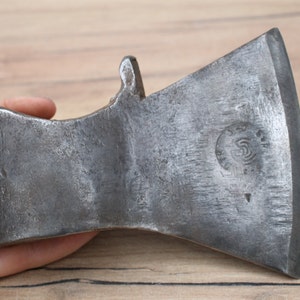 Puede incluir: Una cabeza de hacha grande y resistente con una hoja ligeramente curvada. La cabeza del hacha está hecha de metal y tiene una superficie rugosa y texturizada. La cabeza del hacha tiene una pequeña marca circular en el lateral con un diseño en espiral.