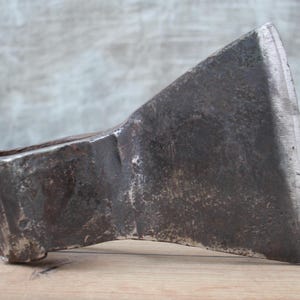 May include: A vintage axe head with a weathered, dark metal finish. The axe head is angled, showcasing the sharp blade and the eye for the handle. The tool rests on a wooden surface, suggesting its use in woodworking or outdoor tasks.