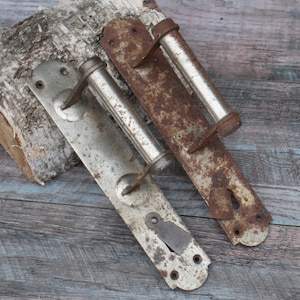 Könnte beinhalten: Zwei antike Türklinken mit silberner Oberfläche und rostigen Akzenten. Die Griffe sind aus Metall und haben ein schlichtes Design. Sie sind an einer Holzoberfläche befestigt.
