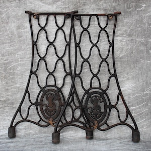 Puede incluir: Dos patas de mesa de hierro fundido negro con un diseño decorativo tejido y un emblema circular con un pájaro estilizado y texto. Las patas son curvadas y tienen una base redondeada.