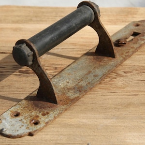 Può includere: Una maniglia per porta in metallo antico con manico nero. La base rettangolare è molto arrugginita, mostrando segni di età e utilizzo. Il manico è cilindrico e sembra essere fatto di un materiale simile alla gomma. La base ha ritagli decorativi.