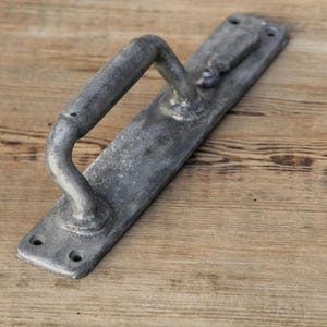 May include: A weathered, gray metal door handle with a curved grip and rectangular backplate. The handle has a vintage appearance, with visible wear and tear, and is mounted on a wooden surface.