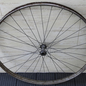 Op de afbeelding: Een antieke fietswiel met een versleten metalen velg en talrijke dunne spaken die uit een centrale naaf stralen. Het wiel heeft een verouderd uiterlijk, met tekenen van roest en slijtage.