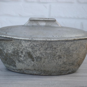 May include: Oval-shaped, vintage aluminium casserole dish with a lid. The dish has a handle on the lid and a textured, aged appearance. The casserole dish is likely used for cooking and serving food.