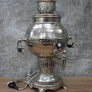 May include: A silver-colored samovar with a rounded body, spigot, and decorative handles. The tea kettle has a cylindrical base and a lid with a decorative top. A power cord with a plug is connected to the base.