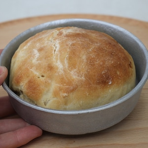 Może przedstawiać: Okrągły bochenek chleba w srebrnej formie do pieczenia. Chleb jest złotobrązowy i ma lekko chrupiącą skórkę.