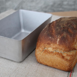 Puede incluir: Una hogaza de pan recién horneado junto a un molde de pan plateado sobre un mantel de lino blanco.