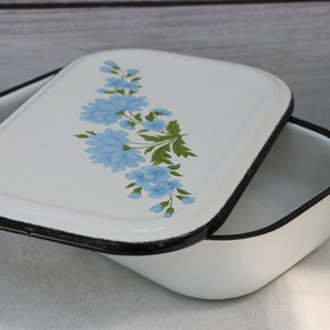 May include: White enamelware baking dish with a black rim and a floral design featuring blue flowers and green leaves on the lid.