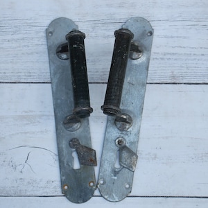 Puede incluir: Dos manillas de puerta vintage con manijas de metal negro y placas de metal plateado. Las manijas están fijadas a las placas con tornillos.