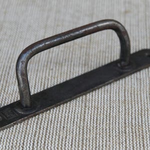 May include: A rustic, dark metal handle with a curved grip and a rectangular base. The handle is attached to the base with rivets. The metal has a weathered appearance, suggesting age and use. Ideal for furniture restoration or industrial decor.