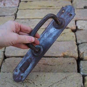 May include: A rustic, iron door handle with a curved handle and a rectangular plate. The handle is weathered and has a dark gray patina.