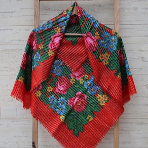 以下が含まれることがあります： 緑色の花柄が施された赤い正方形のスカーフ。花はピンク、青、黄色。スカーフはフリンジの縁取りがされている。