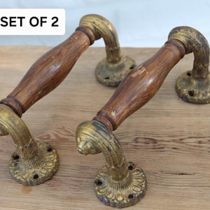 May include: A set of two door handles featuring brown wooden grips and ornate, weathered gold-tone metal mounting plates and ends. Each handle has a curved design with circular bases containing screw holes. The text SET OF 2 is visible.