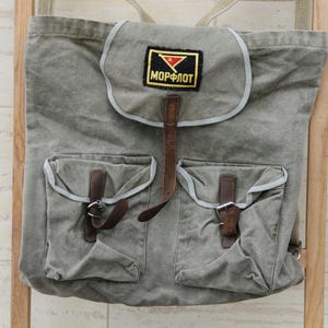 Può includere: Zaino in tela verde oliva in stile vintage con due tasche frontali e cinturini in pelle. Lo zaino presenta una toppa con la scritta "МОРФЛОТ" in lettere nere. La borsa è appesa a un telaio di legno.