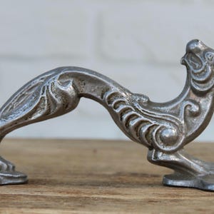 May include: A silver-toned metal door handle shaped like a stylized creature with an arched back and an elongated neck. The handle features intricate embossed scrollwork and textured detailing along its body, with two flat mounting plates at each end.