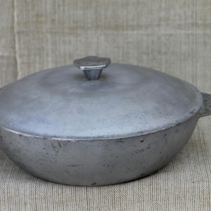 Puede incluir: Una olla redonda de metal color plata con su tapa a juego. La olla cuenta con dos asas laterales y un pomo central en la tapa. La superficie del metal presenta un acabado mate y texturizado con signos visibles de desgaste y pátina.