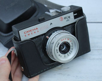 May include: A black and silver 35mm film camera with a leather case. The camera has a lens with a silver ring and the text "СМЕНА SMENA 8M A" on the top. The lens has a red and white scale with numbers and markings.