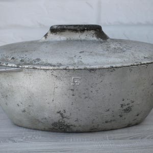 May include: A vintage, oval-shaped metal cooking pot with a lid. The pot is a dull silver colour with signs of wear and age, including dark spots and discolouration. The lid has a small handle and a lip for easy removal. A small, stylized letter "S" is visible on the side.