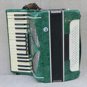 May include: A green marbled accordion featuring a white keyboard with black keys, a central silver metal grille, and a side panel with white circular buttons. The instrument includes a black bellows section and a small circular logo with a harp icon.