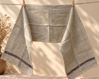 Puede incluir: Un camino de mesa rectangular de tela color beige claro y textura gruesa colgado de un tendedero. La tela presenta una banda decorativa horizontal cerca de cada extremo, con un patrón geométrico repetitivo en negro y rosa.