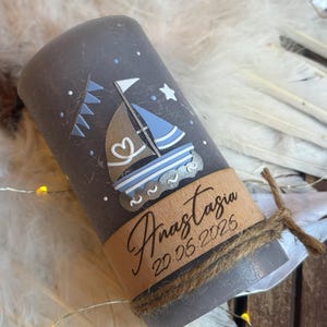 Könnte beinhalten: Eine graue Säulenkerze mit einem Segelboot-Design in Braun und Blau, mit einem Herzdetail. Die Kerze hat ein braunes Band mit dem Namen "Anastasia" und dem Datum "20.06.2026" aufgedruckt. Eine Schnur ist um das Band gewickelt.