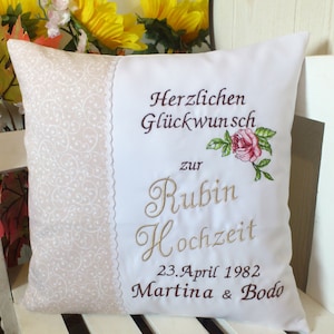 Può includere: Cuscino quadrato bianco con testo ricamato e motivo floreale. Il cuscino presenta le parole "Herzlichen Glückwunsch zur Rubin Hochzeit" e la data "23. April 1982". Una rosa rosa è ricamata sul cuscino.