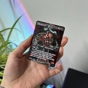 Puede incluir: Una carta coleccionable con el texto "Massacre Girl". Presenta un personaje femenino con armadura roja y negra. La carta se sostiene en una mano.