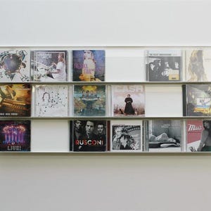 Op de afbeelding: Een aan de muur gemonteerde cd-display met verschillende muziekalbums. De albums hebben diverse albumhoezen, waaronder bandfoto's, illustraties en tekst. De unit is wit en gemonteerd op een witte muur.