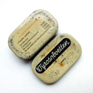 May include: Two small, rectangular, metal tins with white lids. The top tin has the text "Zusammensetzung" and a list of ingredients in German. The bottom tin has the text "Tyrosolvetten" in black lettering on a white background.