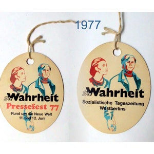 Könnte beinhalten: Zwei runde, cremefarbene Anhänger mit einer Schnur. Der Anhänger links zeigt eine rote, blaue und schwarze Grafik von zwei Personen und den Text "die Wahrheit Pressefest 77 Rund um die Neue Welt 11. und 12. Juni". Der Anhänger rechts zeigt eine rote, blaue und schwarze Grafik von zwei Personen und den Text "die Wahrheit Sozialistische Tageszeitung Westberlins".